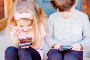 Zwei Kinder, ein Junge und ein Mädchen, sitzen nebeneinander und haben jeweils ein Handy in der Hand. Ihre Blicke liegen auf den Mobilgeräten vor ihnen.